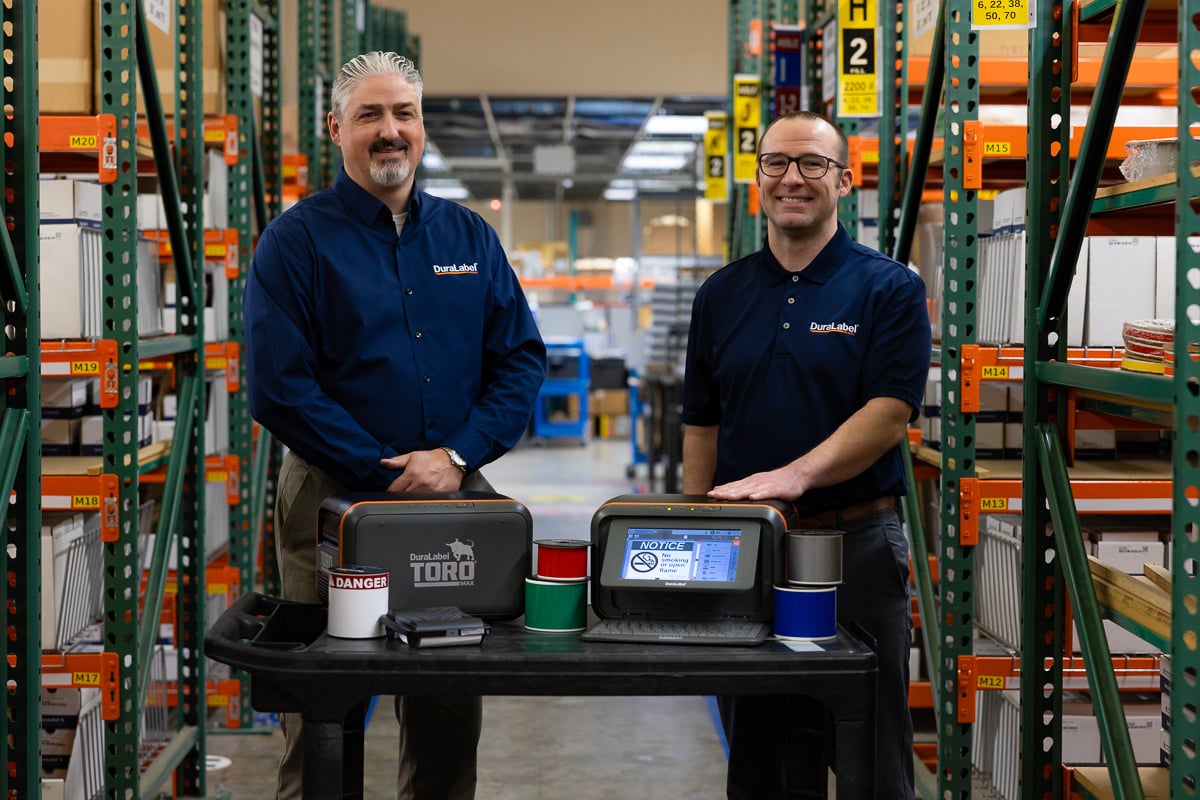  account-managers-joshua-day-and-joe-wolf-standing-in-front-of-the-kodiak-max-and-toro-max-industrial-label-and-sign-print-systems-at-the-duralabel-warehouse